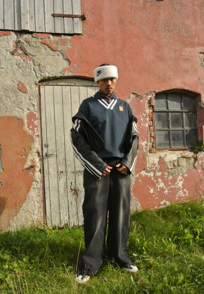 Μπλε αθλητικό μπλουζάκι με άσπρες ρίγες, μαύρο μπουφάν με ριγωτά μανίκια, φαρδιά σκούρα τζιν και άσπρη κορδέλα στο κεφάλι, που στέκεται μπροστά από έναν υφαντό τοίχο.