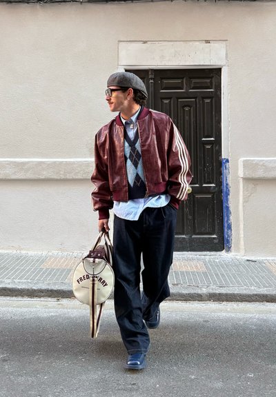 Hombre con chaqueta bomber roja y suéter de rombos, sosteniendo un bolso redondo de Fred Perry, caminando por una acera de la ciudad frente a una puerta negra.