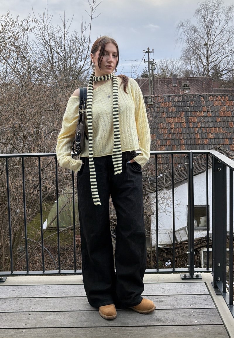 Jeune femme sur un balcon portant un pull jaune à épaules dénudées, une écharpe rayée, un pantalon large noir, des bottes couleur camel, et tenant un sac à main noir.