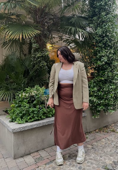 Americana de cuadros beige sobre un top blanco corto, falda de satén bronce y zapatillas ligeras; rodeada de plantas verdes y pavimento de piedra.