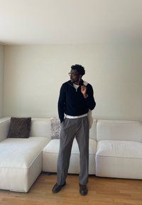 Mężczyzna w okularach, ubrany w czarny sweter i szare spodnie, stoi w salonie z białą, modułową sofą i wzorzystymi poduszkami.