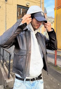 Fekete bőrkabát rövidített fazonnal és két első zsebbel, szürke kapucnis pulóver fölött viselve, világoskék farmerral és sapkával kombinálva.