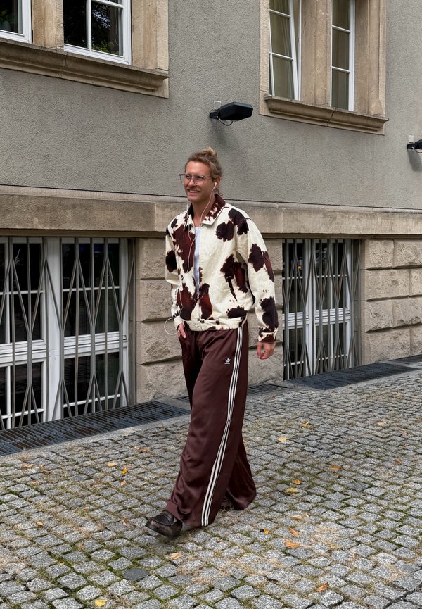 Persona con chaqueta con estampado y pantalones deportivos marrones camina por una calle adoquinada junto a un edificio con ventanas con rejas y paredes grises.