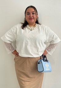 Λευκό μπλουζάκι με δαντελένια μανίκια, μπεζ φούστα και μικρή μπλε λαμπερή τσάντα Adidas με μαύρες λεπτομέρειες και λογότυπο. Μεγάλο λευκό κολιέ με μαργαρίτες.