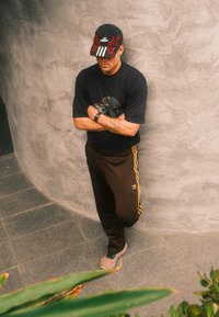 Black T-shirt with graphic print, brown adidas track trousers with gold stripes, and a cap featuring a red flame design and three stripes.