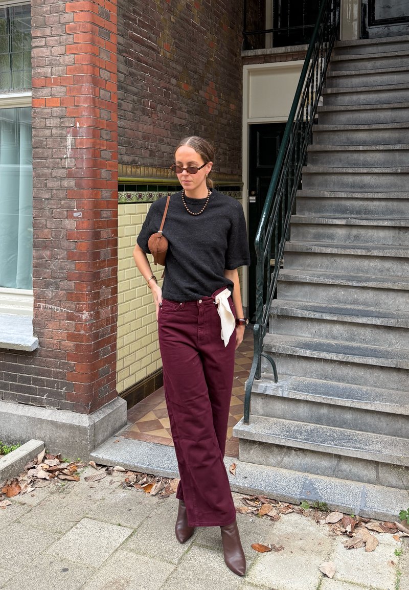 Pull noir à manches courtes, pantalon bordeaux, bottines marron. Accessoires : petit sac à bandoulière marron et collier en perles. Fond urbain.