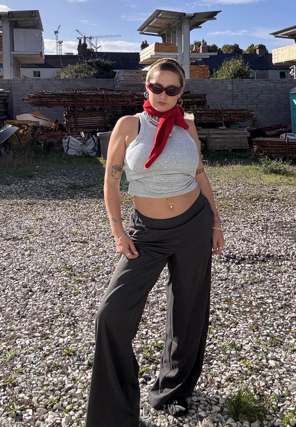Une femme portant des lunettes de soleil, une écharpe rouge et un pantalon sombre ample se tient sur du gravier dans un chantier.