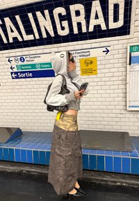 Grijze cropped hoodie met capuchon, camouflage maxi rok met zakken en zwarte puntige ballerina's, voor een achtergrond van een metrostation.