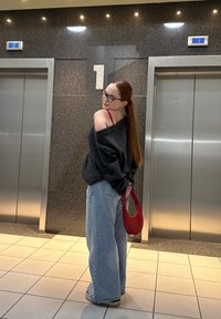 Oversized donkergrijze trui, lichtblauwe jeans met wijde pijpen, rode tas en bril. Gladde texturen in een moderne liftomgeving.