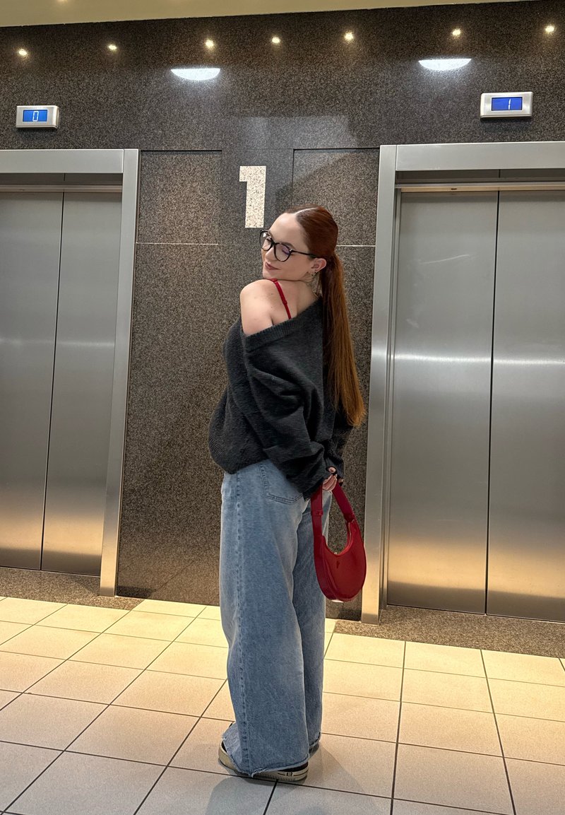 Pull oversize gris foncé, jeans larges bleu clair, sac rouge et lunettes. Textures lisses dans un cadre moderne d'ascenseur.
