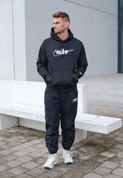 Hombre con sudadera con capucha y pantalones negros de Nike, zapatillas blancas, de pie con las manos en los bolsillos cerca de un banco de concreto blanco al aire libre.