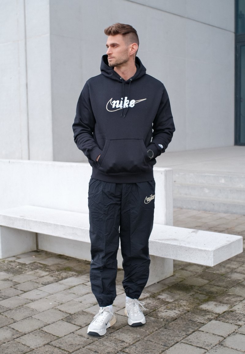 Homme portant un sweat à capuche et un pantalon Nike noirs, des baskets blanches, debout les mains dans les poches près d'un banc en béton blanc en plein air.