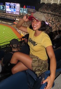 Gul T-skjorte med svart grafikk, kamuflasje skjørt og en beige caps med rød brem. Sitter i stadionseter med et resultatbrett i bakgrunnen.