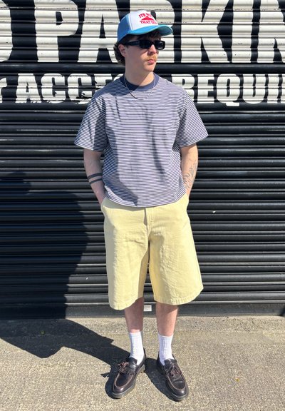 Camisa de manga corta a rayas en azul marino y blanco, pantalones cortos amarillos hasta la rodilla, zapatos negros, calcetines blancos y una gorra azul con texto.