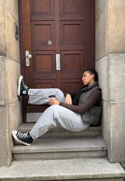 Una mujer se sienta en los escalones de piedra junto a una puerta de madera, vistiendo un chaleco acolchado negro, una camiseta de manga larga marrón, unos Jeans gris holgados y zapatillas negras. Sostiene una taza de café.