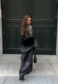 Γυναίκα με μακριά μαλλιά, ντυμένη με ρούχα με λεοπάρ σχέδιο, κρατώντας ένα μαύρο δερμάτινο παλτό με γούνα, στέκεται σε λιθόστρωτο πεζοδρόμιο μπροστά από μια σκούρα πράσινη πόρτα.