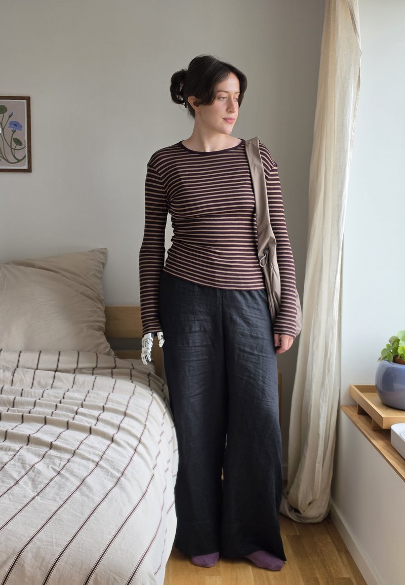 Young woman with a prosthetic left hand stands next to a bed wearing a striped shirt, black pants, and purple socks in a bright bedroom.