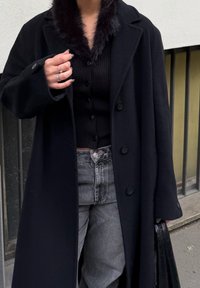 Manteau long noir avec un col en fausse fourrure, associé à un haut côtelé noir boutonné et un jean taille haute gris foncé. Tient un sac noir.
