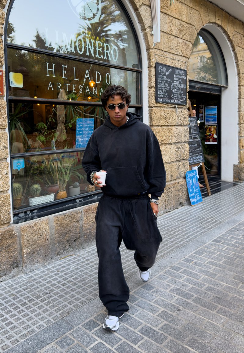 Hoodie noir surdimensionné, pantalon large assorti, baskets blanches et lunettes de soleil rondes. Tenant un gobelet jetable, marchant sur un pavé en pierre.