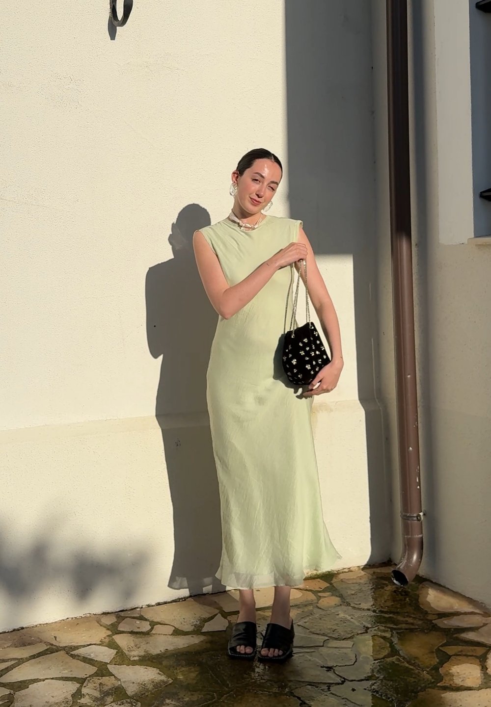 Light green, sleeveless dress with a high neck; sheer fabric. Black velvet handbag with a white floral design; black slide sandals.