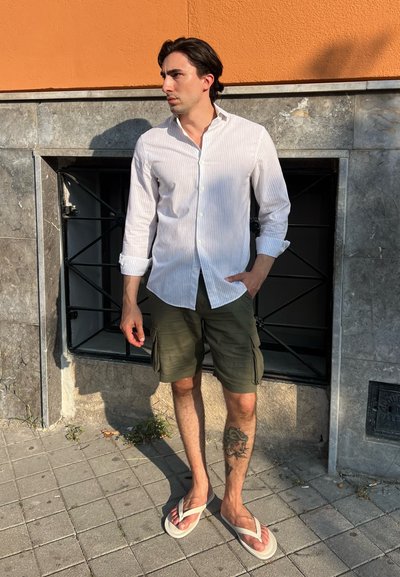 Hombre con una camisa de rayas blanca de manga larga, pantalones cortos de cargo verde oliva y chanclas; de pie contra una pared de piedra.
