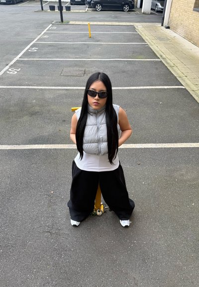 Chaleco tipo puffer en gris claro, camiseta blanca ajustada, pantalones anchos negros, gafas de sol negras, de pie en un aparcamiento con concreto y un bolardo amarillo.