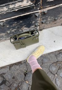 Olivengrønn strukturert håndveske med doble håndtak og metallbeslag, plassert på en marmoroverflate ved siden av en fot i gule joggesko og rosa sokker.