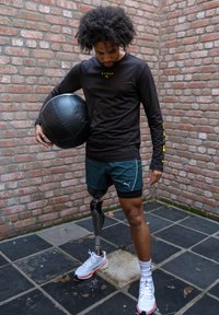 Must pikkade varrukatega särk kollase logoga, rohelised lühikesed püksid ja valged spordijalatsid. Isik hoiab musta meditsiinipalli; tal on proteesjalg.