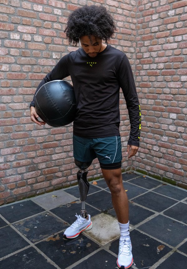 Camiseta preta de manga comprida com logótipo amarelo, calções verdes e sapatos atléticos brancos. A pessoa segura uma bola medicinal preta; tem uma perna protética.
