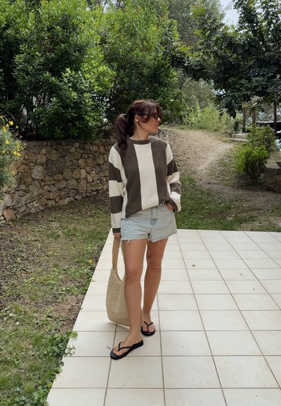 Suéter a rayas marrón y crema, pantalones cortos de denim claro y chanclas negras. Sosteniendo una bolsa de mimbre, de pie sobre un pavimento de baldosas con vegetación.