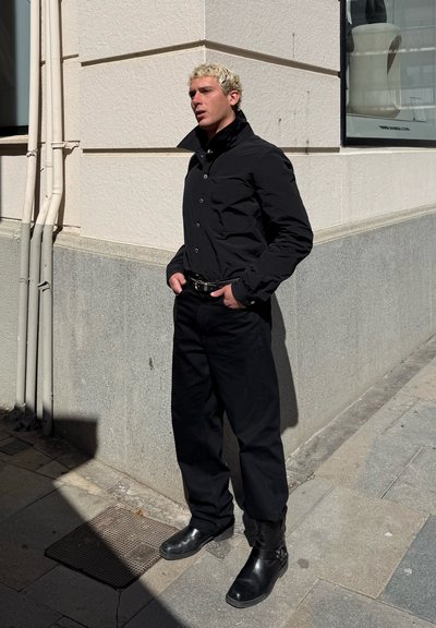 Camisa negra de botones con cuello alto, combinada con pantalones negros holgados y botas de tobillo de cuero negro. Pared de cemento y luz solar de fondo.