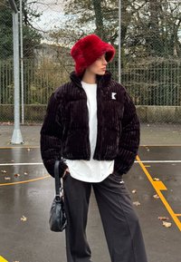 Personne portant un chapeau seau rouge pelucheux, une doudoune noire, une chemise blanche et un pantalon noir, debout sur un terrain de basket en plein air.