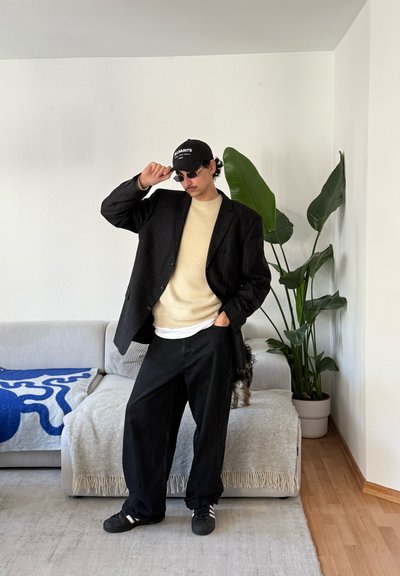 Hombre con blazer negro de rayas, suéter beige, pantalones anchos negros, gorra y gafas de sol, posando en una sala de estar con sofá y una planta grande y verde.