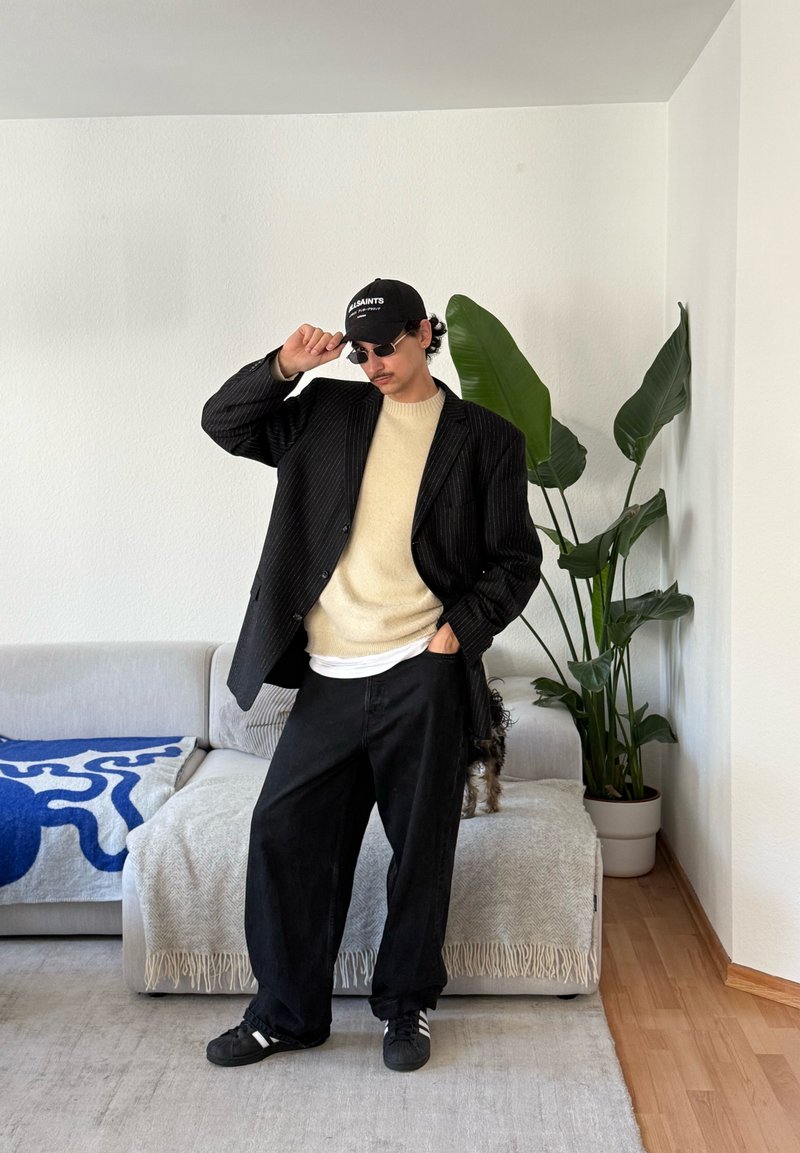 Homme portant un blazer noir à fines rayures, un pull beige, un pantalon large noir, une casquette et des lunettes de soleil, debout dans un salon avec un canapé et une grande plante verte.