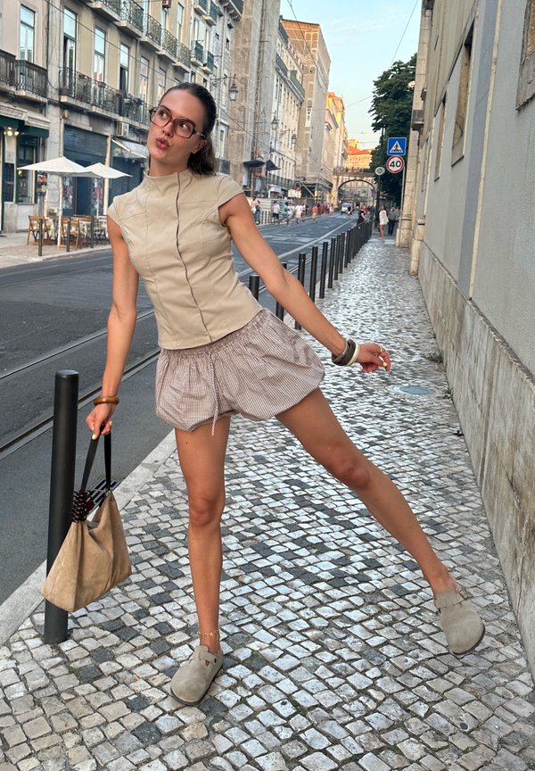 Jeune femme portant des lunettes, un haut beige et un short, posant de manière espiègle sur un trottoir pavé dans une rue de la ville.