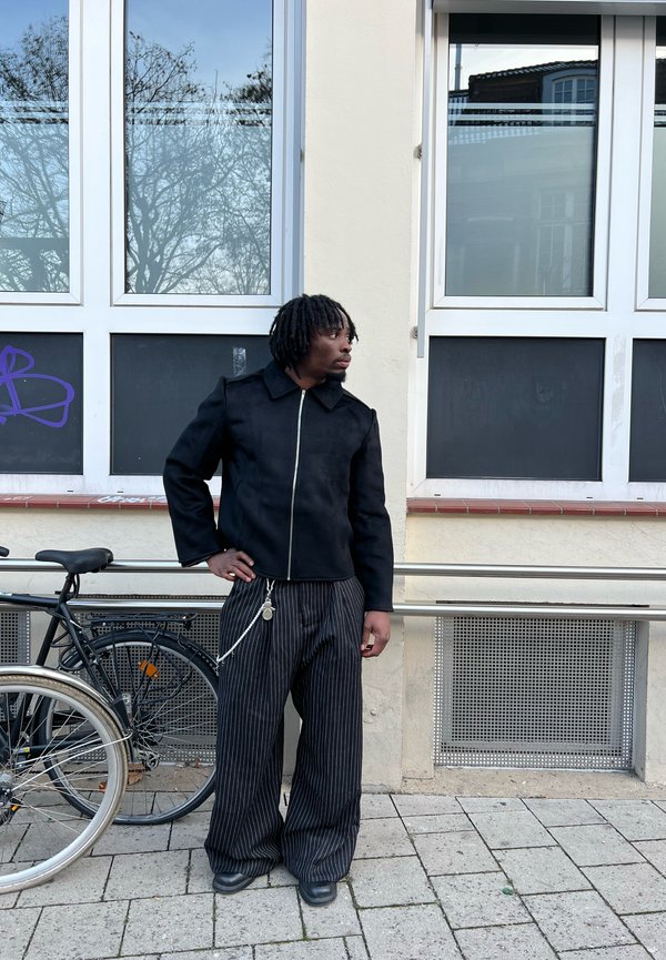 Man wearing a black jacket and wide pinstripe trousers standing on the pavement next to two bicycles, looking to the right in an urban setting.