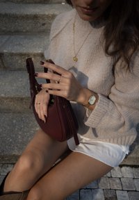 Bordeaux læder håndtaske med guld hardware, struktureret overflade, holdt i hænderne med en beige sweater, guld halskæde og et ur på håndleddet.