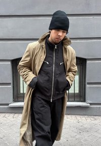 Νεαρός άνδρας που φοράει μαύρο σκουφί, μαύρο μπουφάν με φερμουάρ και μπεζ παλτό περπατά σε πεζοδρόμιο της πόλης με τα χέρια στις τσέπες.