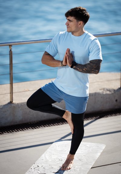 Camiseta deportiva azul claro, mallas negras y pantalones cortos azules. La persona está de pie sobre una pierna en una postura de yoga sobre una esterilla con textura.