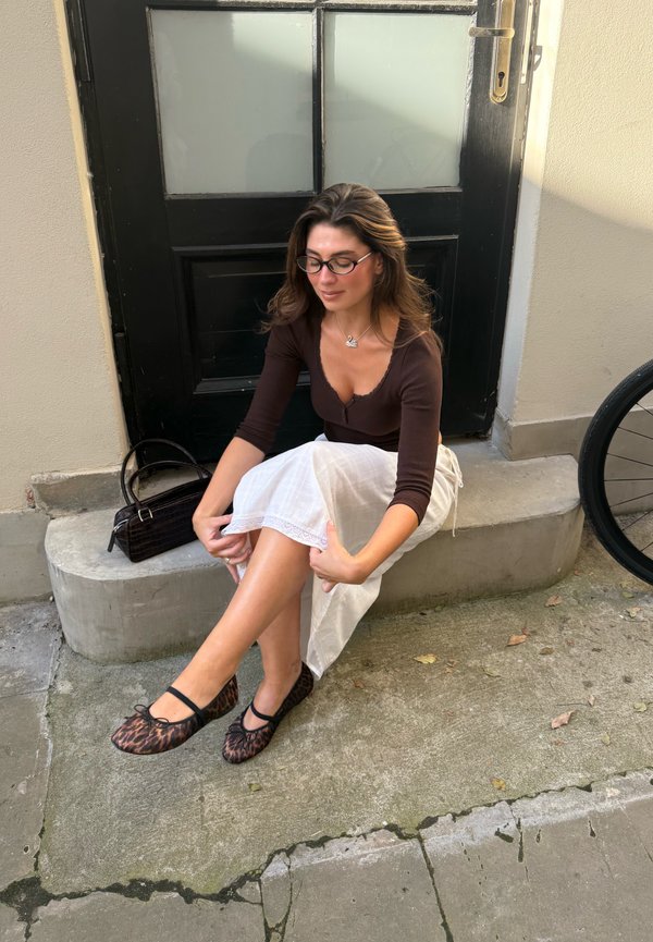 Mujer con gafas y una blusa marrón ajusta una falda blanca mientras está sentada en unas escaleras de hormigón al aire libre junto a una puerta negra, con un bolso cerca.