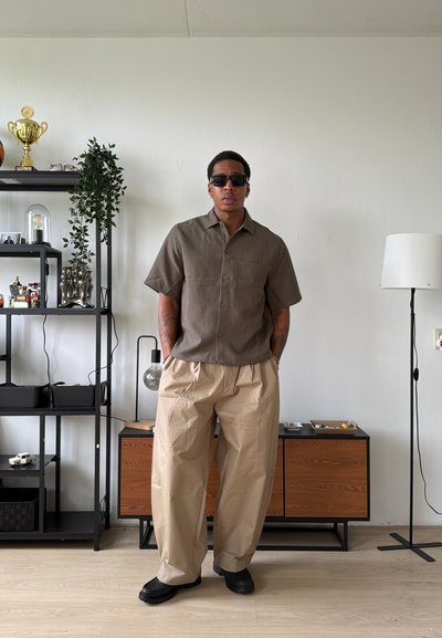 Hombre con gafas de sol negras, camisa marrón de manga corta y pantalones anchos beige, de pie en una habitación moderna con estanterías y una lámpara de pie.