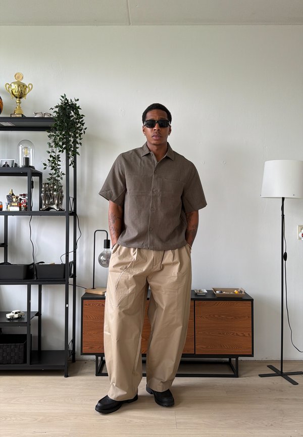 Homme portant des lunettes de soleil noires, une chemise marron à manches courtes et un pantalon beige large, debout dans une pièce moderne avec des étagères et une lampe sur pied.