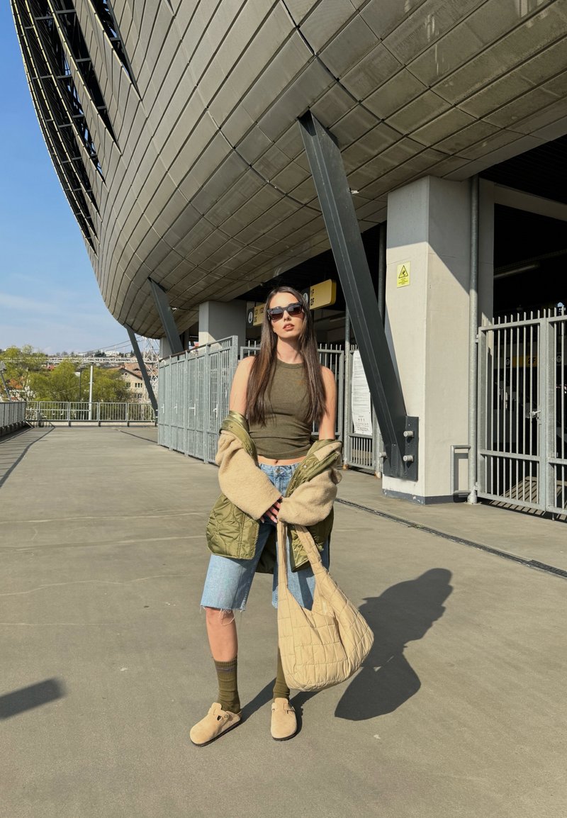 Ελαιοπράσινο αμάνικο μπλουζάκι, υπερμεγέθη πράσινη πουκαμίσα με επένδυση, τζιν σορτσάκι, καφέ τσάντα ώμου, ελαιοπράσινες κάλτσες και μπεζ παπούτσια slip-on μπροστά από φόντο σταδίου.