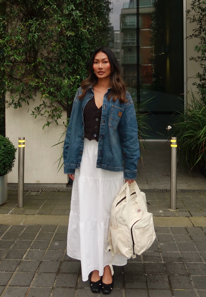 Veste en denim de teinte bleue, haut noir à boutons, jupe blanche à volants et sac à dos crème. Chaussures noires et verdure en arrière-plan.