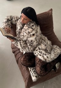 Faux fur-kappa med svart och vit prickig mönster, bärs över spetsstrumpbyxor, med en brun översizekudde och en bok i handen.