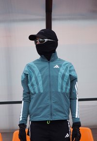 Veste de sport bleu sarcelle avec des accents noirs et des rayures blanches, associée à des gants noirs, une casquette noire et des lunettes de soleil.
