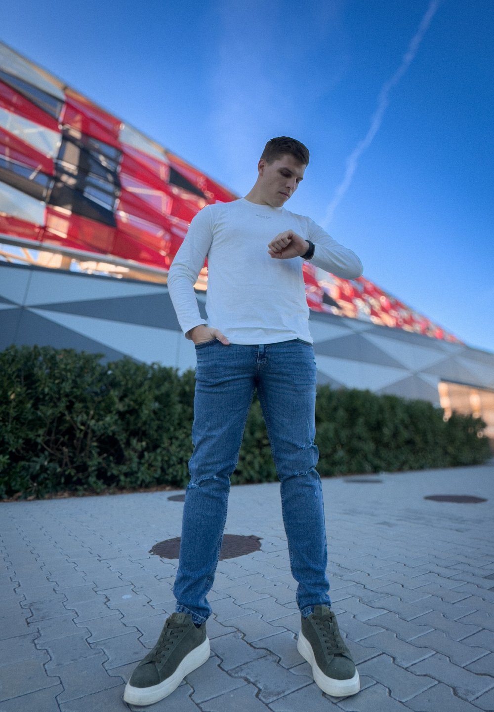 Λευκό πουκάμισο με μακριά μανίκια, μπλε τζιν με φθορές και πράσινα αθλητικά παπούτσια ψηλά στον αστράγαλο. Άτομο που ελέγχει το ρολόι του μπροστά από σύγχρονο κτήριο.