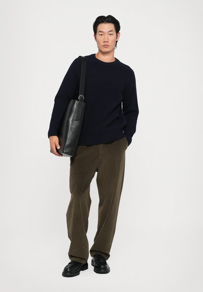 Suéter de canalé azul marino, pantalones de pana oliva, bolso de cuero negro con correa para el hombro, zapatos negros, de pie contra un fondo blanco liso.