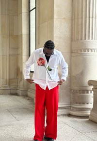 Chemise blanche boutonnée avec imprimé de rose rouge, associée à un pantalon rouge ample. Chaussures noires et lunettes complètent le look. Texture de tissu lisse.