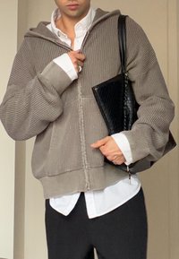 Grijze ribgebreide hoodie met een rits aan de voorkant, oversized mouwen en een wit overhemd met kraag eronder, gecombineerd met een zwarte tas met structuur.